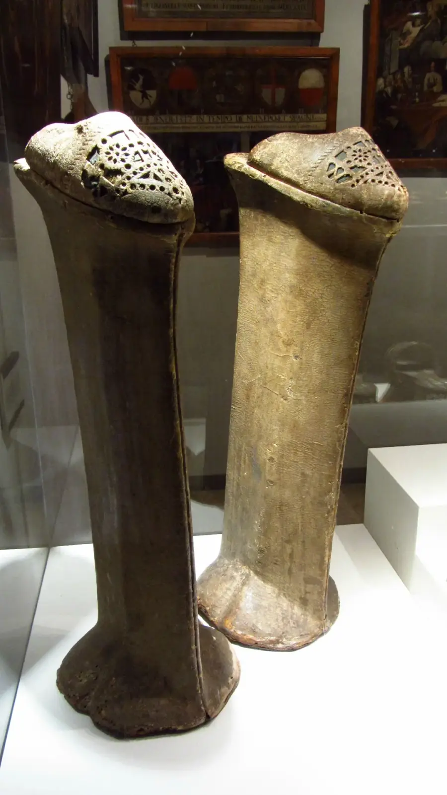 Two tall, curved, brown shields with rounded tops and perforated designs are displayed upright in a museum exhibit case. The shields have a textured, worn surface and sit on a white platform.