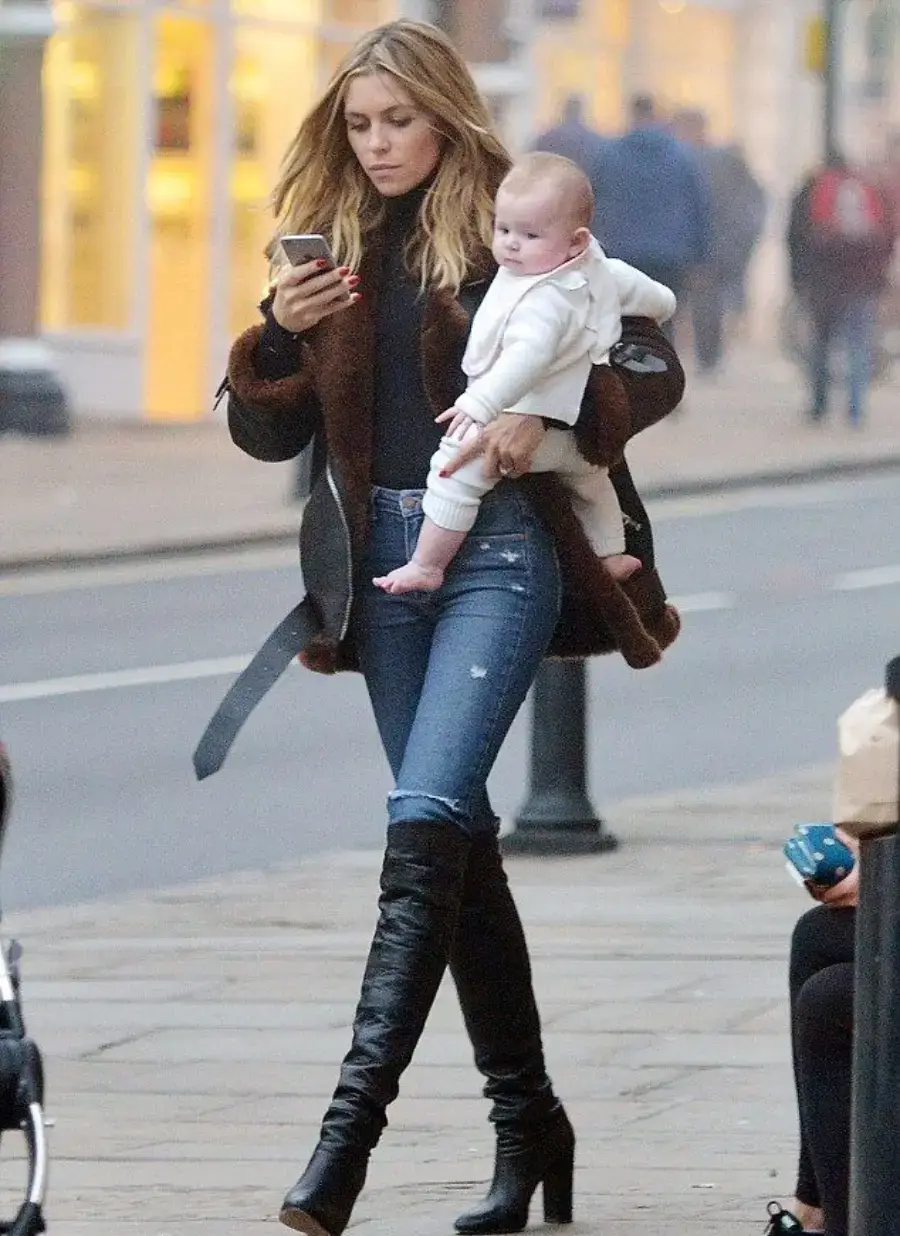 A woman in a black coat, jeans, and heeled boots walks on a sidewalk, holding a baby in one arm and looking at her phone with the other hand.