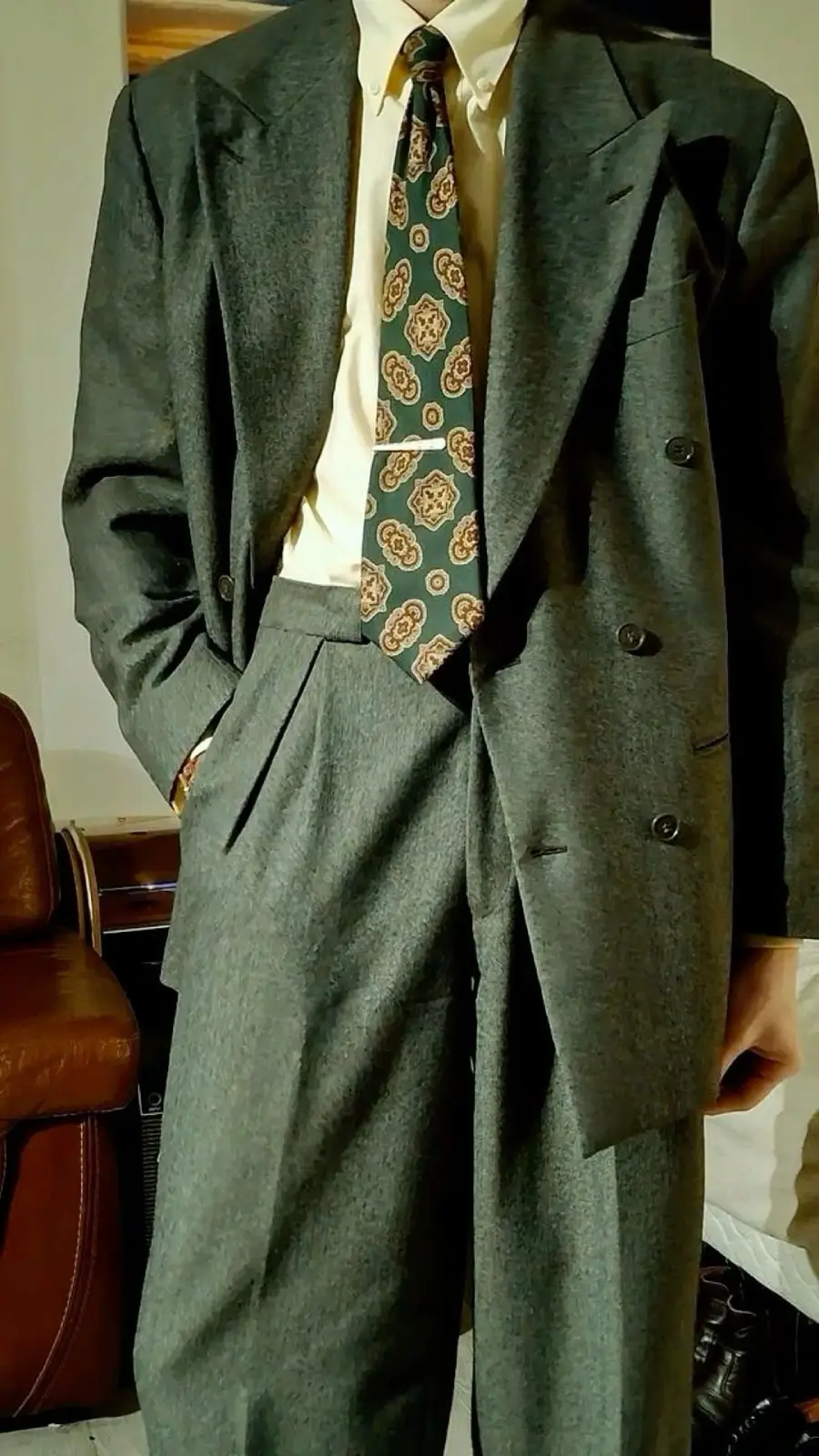 Person wearing a loose-fitting gray suit, cream shirt, and green patterned tie, with one hand in their pocket, standing indoors near a brown leather chair.