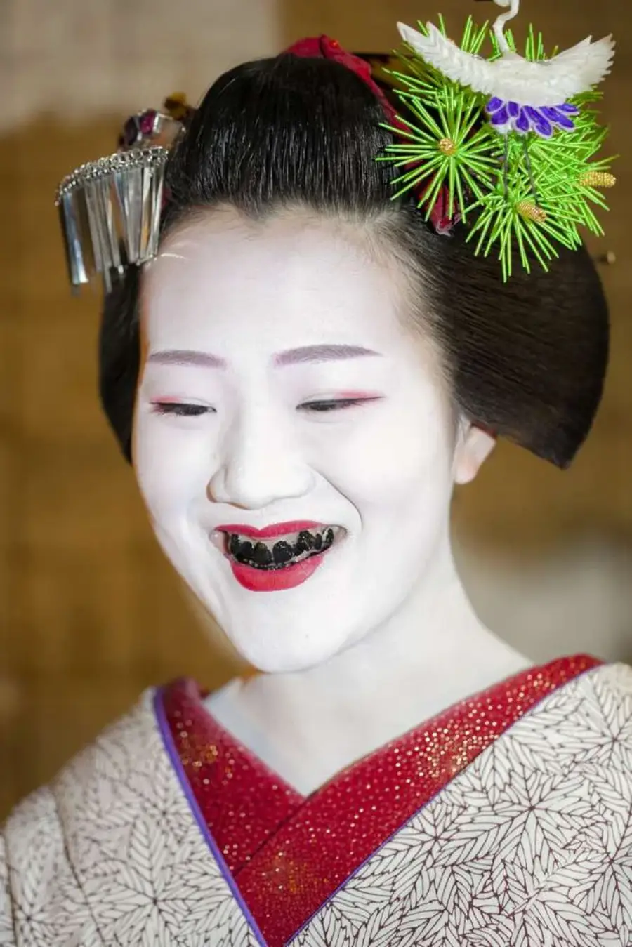 A woman dressed as a geisha with white face makeup, blackened teeth, red lipstick, and elaborate hair ornaments, wearing a patterned kimono with a red collar.