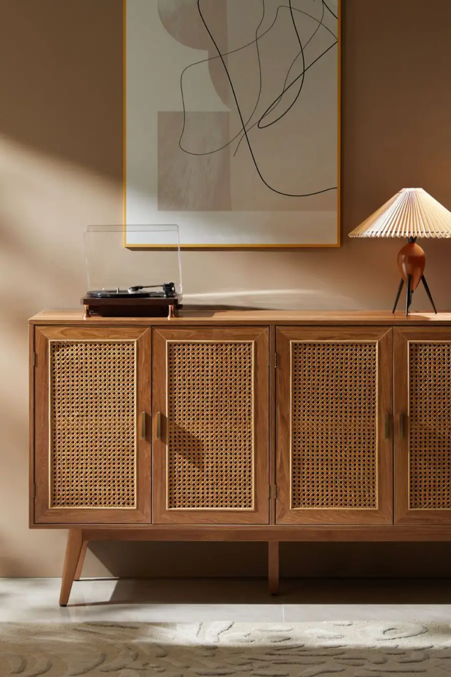 A mid-century modern wood sideboard with rattan doors holds a turntable and a lamp. Above it hangs an abstract line art piece. The setting features neutral walls and a patterned light rug.