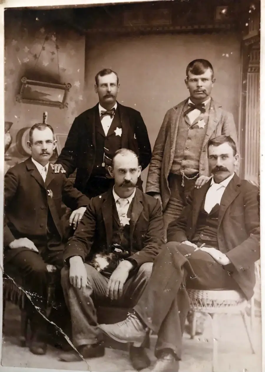 Five men in late 19th-century suits pose for a formal portrait in a vintage studio with ornate decor; three are seated in front and two stand behind them, all with serious expressions and mustaches.