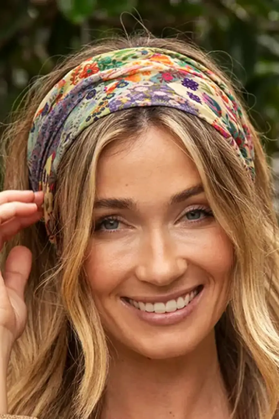 Smiling woman with long blonde hair wearing a colorful floral headband, standing outdoors with greenery in the background.