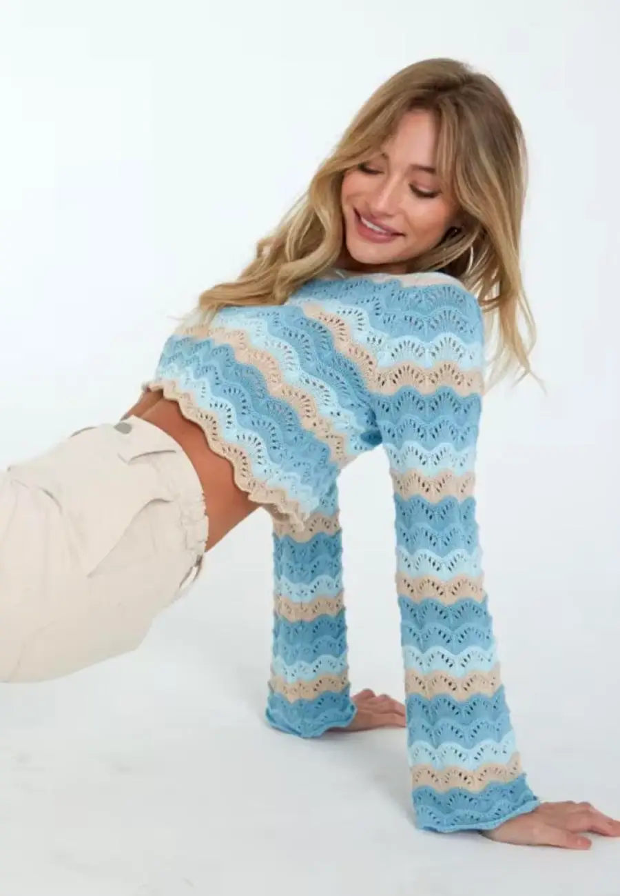 Woman in a blue, white, and beige wavy-striped crochet sweater and beige pants sits on the floor, smiling with eyes closed and hands behind her, against a plain background.