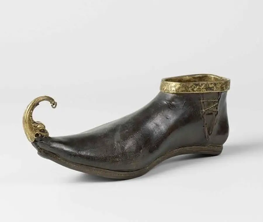 A dark leather medieval shoe with a curved, ornate gold toe and gold trim around the ankle, displayed on a plain, light background.