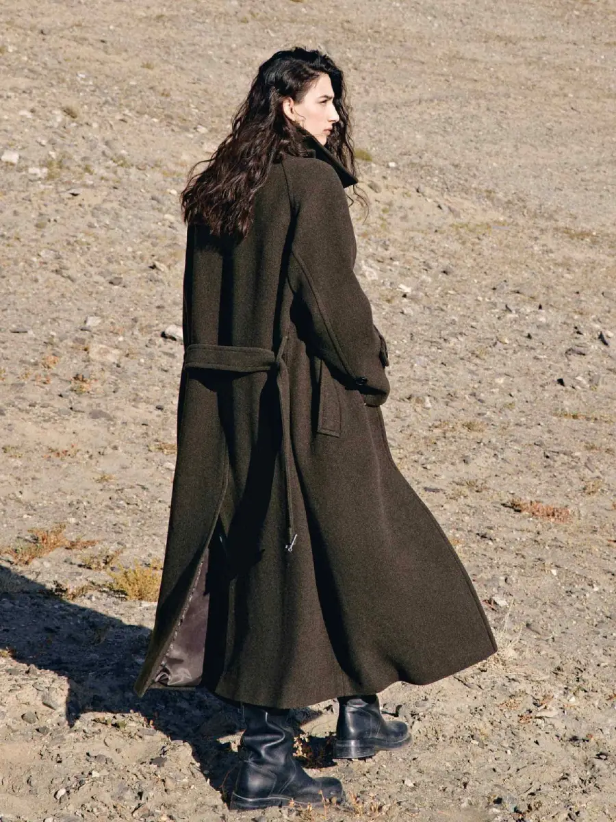 A person with long dark hair stands on rocky ground, wearing a long dark coat and black boots, facing away with their face in profile.