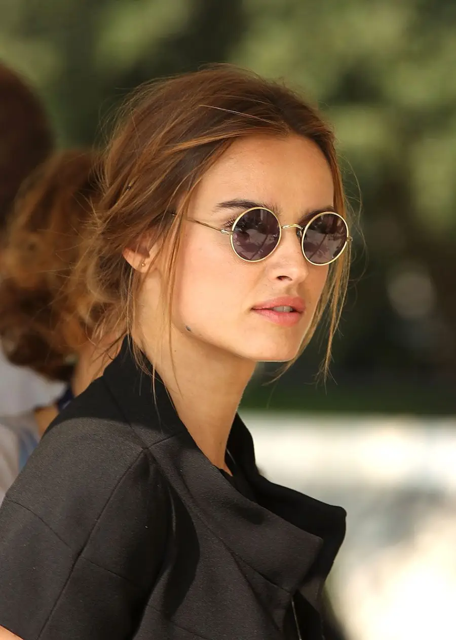 A woman with brown hair in a messy bun wears round sunglasses and a black top, looking to the side outdoors with a blurred background.