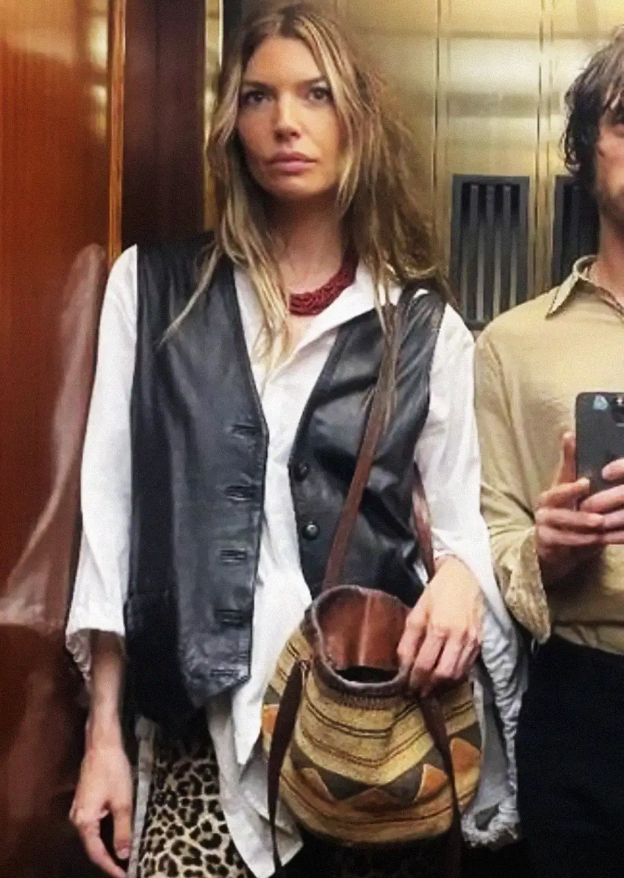 A woman with long hair wears a black leather vest over a white shirt, leopard print pants, a red necklace, and carries a patterned bag. She stands in an elevator next to a man holding a phone.