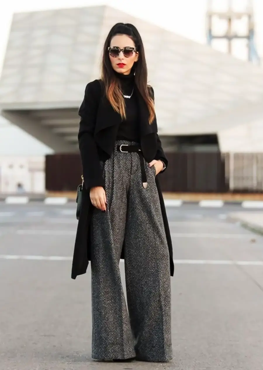 Woman wearing sunglasses, a long black coat, black turtleneck, and wide-leg gray pants stands confidently on a city street with modern architecture in the background.