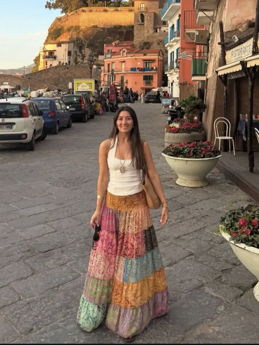 A woman in a colorful patchwork skirt and white tank top smiles while standing on a charming cobblestone street lined with flowers, cars, and colorful buildings.