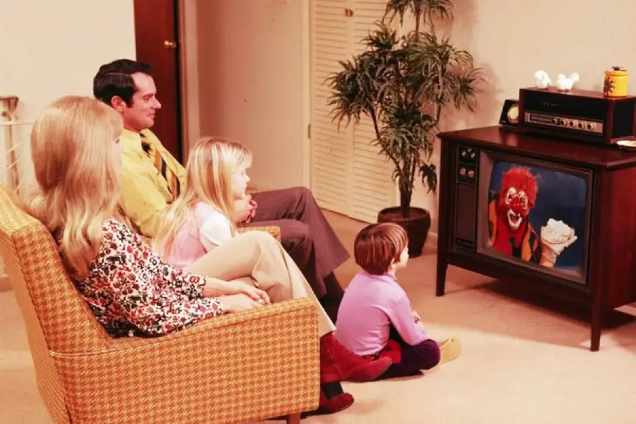 A family of four sits in a living room watching TV, where a clown holding a pie appears on the screen. They sit on a couch and the floor, facing the television. A plant and stereo are visible near the TV.