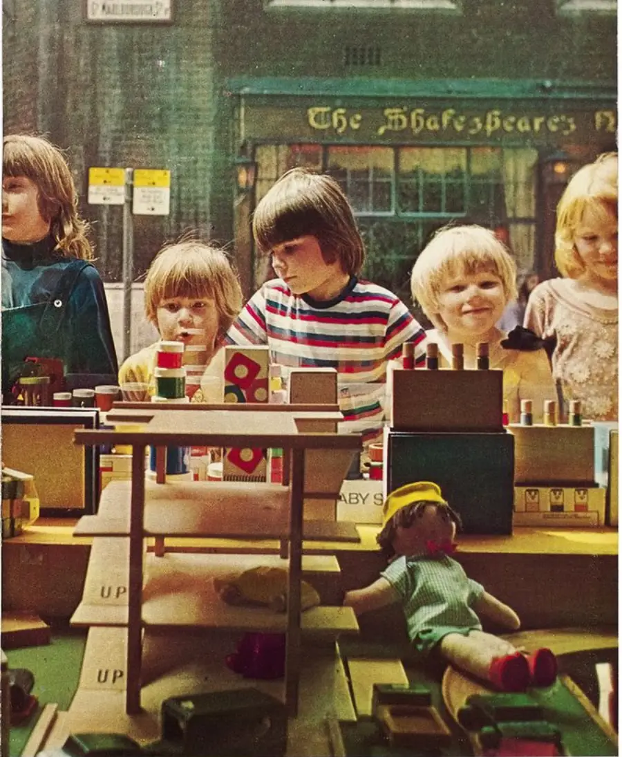 Four children stand behind a toy store window display, looking at wooden toys and a doll. A pub called “The Shakespeare’s” is visible in the background across the street.