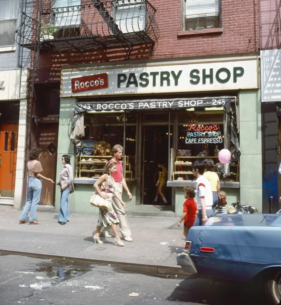 People walk past Rocco’s Pastry Shop on a sunny day; the shop has a green exterior, glass display windows with pastries, and a sign above reading “Rocco’s Pastry Shop.” A blue car is parked in front.