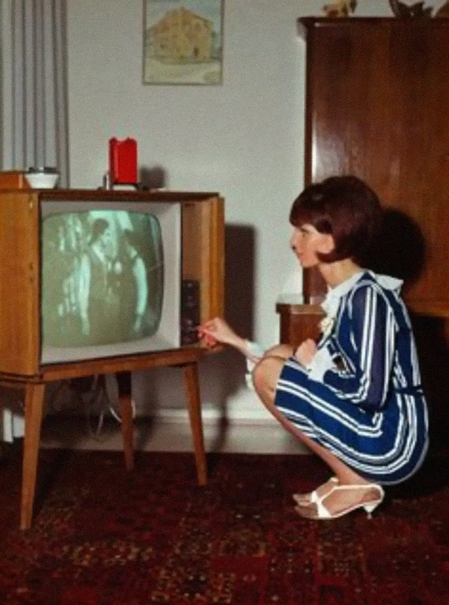 A woman in a blue and white dress kneels to adjust an old-fashioned television set showing a black-and-white program in a vintage living room with a patterned carpet and wooden furniture.