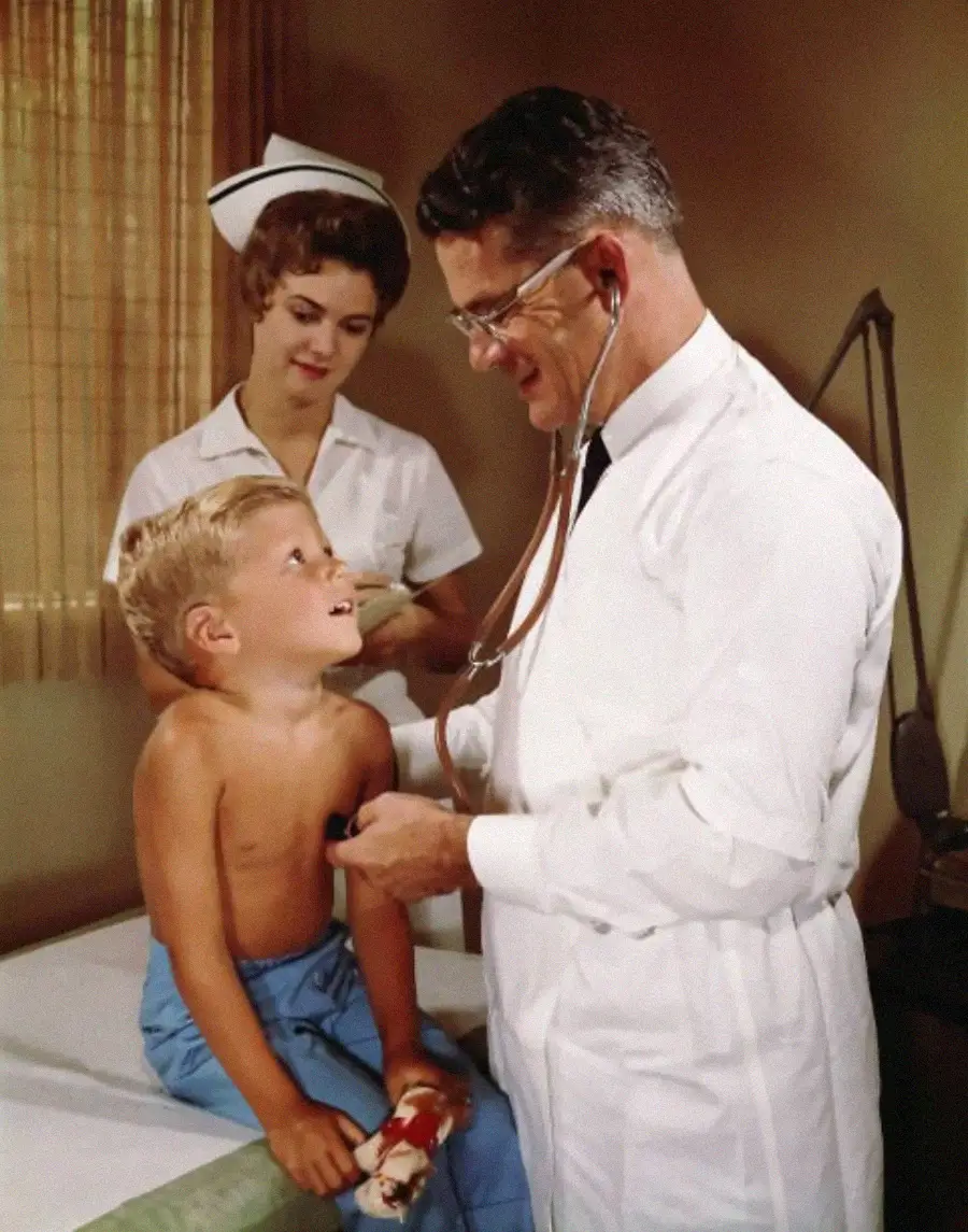 A young boy sits on an exam table as a doctor uses a stethoscope on his chest. A nurse stands nearby, smiling. The boy looks up at the doctor, and medical equipment is visible in the background.