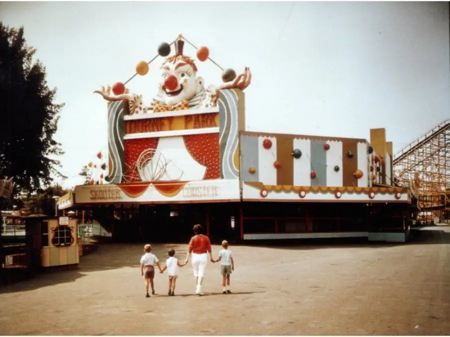 A woman and three children walk toward a large, colorful funhouse with a clown face facade at an amusement park on a sunny day.