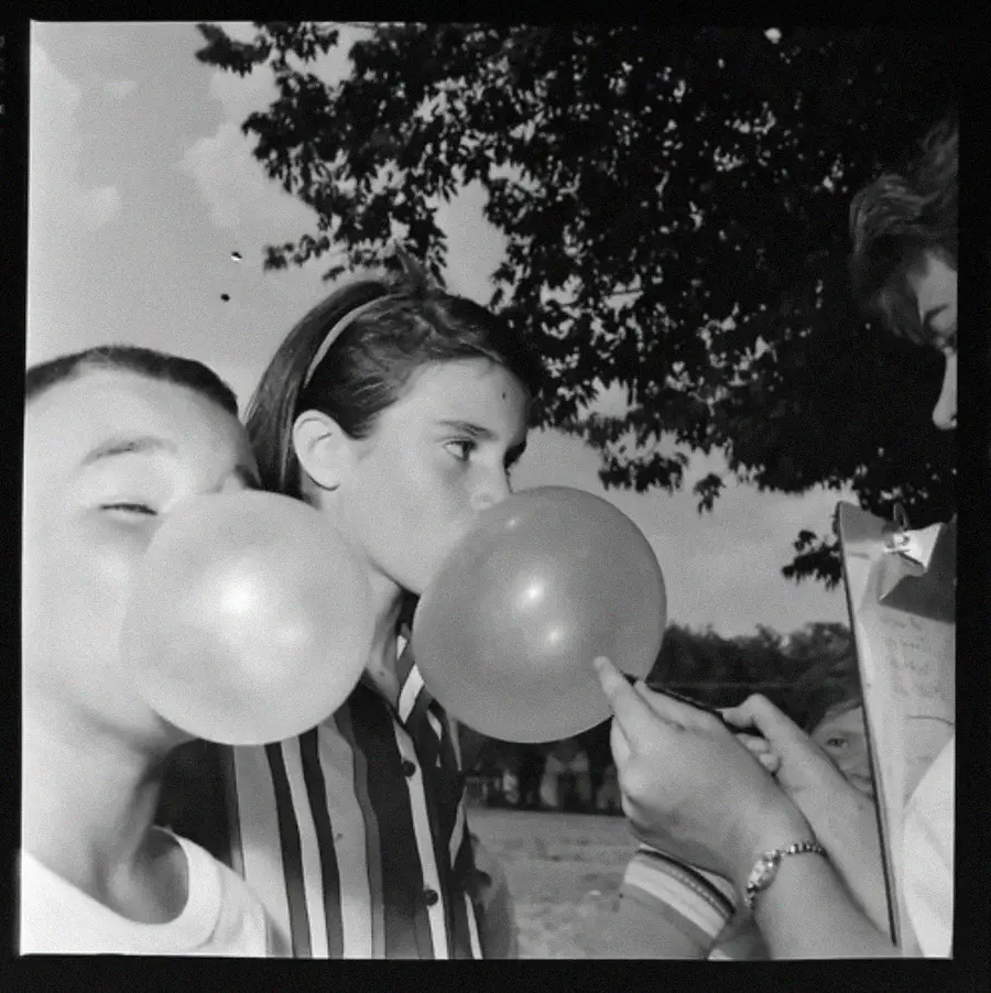 Two children outdoors blow large bubbles with gum as a person with a clipboard measures the size of one bubble. A tree and partly cloudy sky are visible in the background.