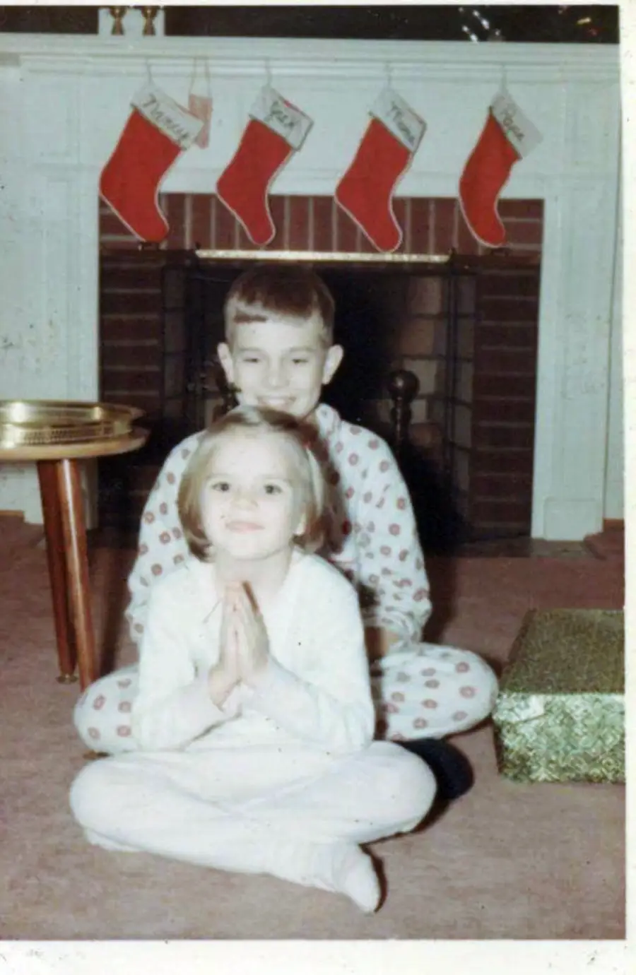 Two children in pajamas sit on the floor in front of a fireplace with four Christmas stockings hanging above. The girl sits cross-legged in front, hands clasped, while the boy sits behind her, smiling. A wrapped present is nearby.