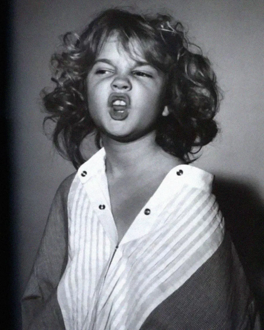 Black and white photo of a young child with curly hair making a playful, exaggerated face, mouth open, and head tilted slightly back. The child wears a striped shirt with the collar loose and buttons undone.