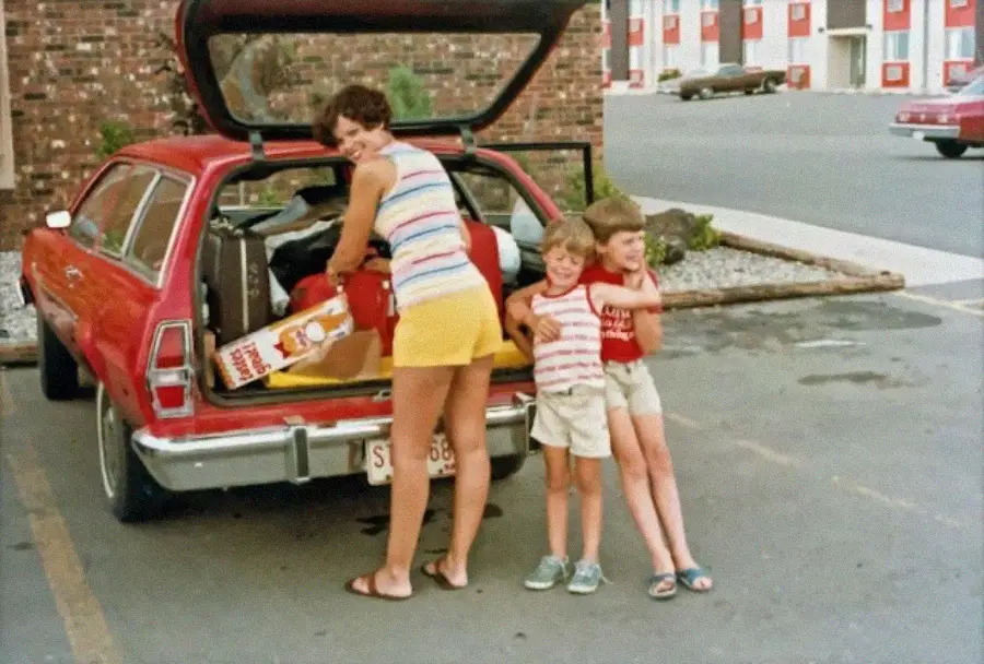 A woman stands by an open red station wagon, packing items, with two smiling young children beside her in a parking lot near a motel.