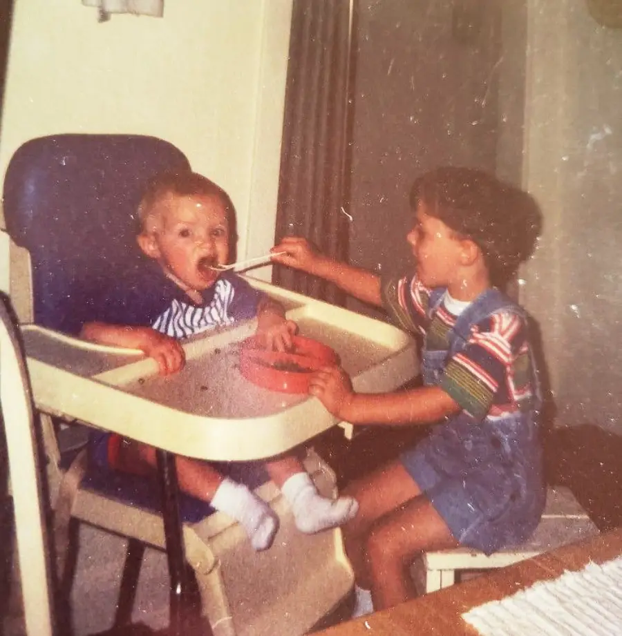 A young child in a high chair is being fed by an older child sitting on a chair. The older child holds a spoon to the younger child’s mouth. Both appear to be indoors at a table.
