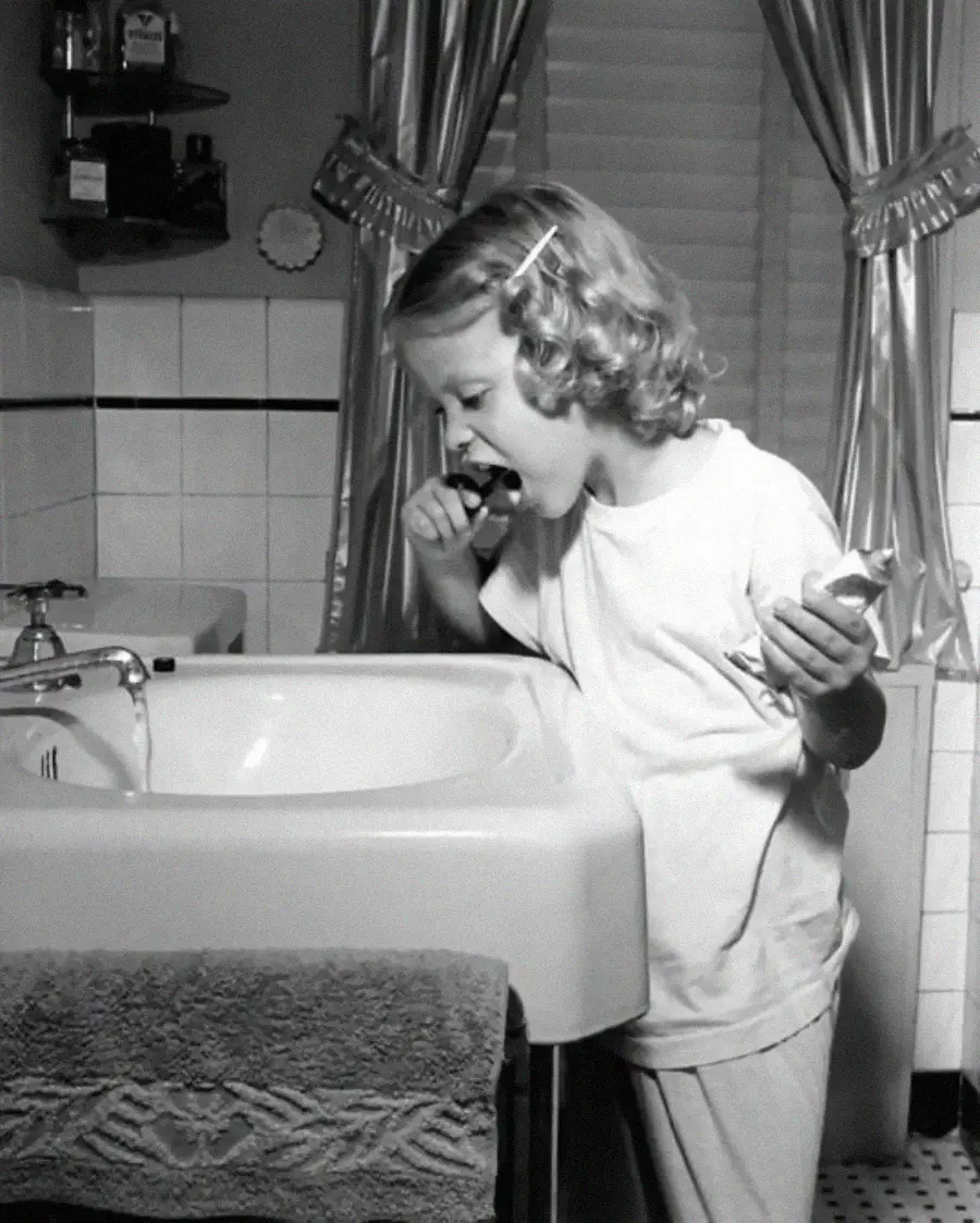 A young child with curly hair brushes their teeth at a bathroom sink, holding a tube of toothpaste and wearing pajamas. The bathroom has tiled walls, a towel, and curtains over the window.