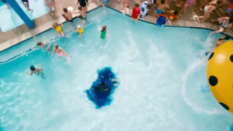 Aerial view of a crowded indoor swimming pool, with children and adults in and around the water. At the pool’s center, blue dye spreads in swirling patterns, while a large, yellow, spotted ball floats nearby.