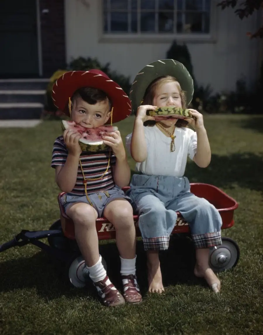 Two young children sitting in a red wagon on grass, wearing colorful hats and eating watermelon slices, smiling and enjoying a sunny day outside.