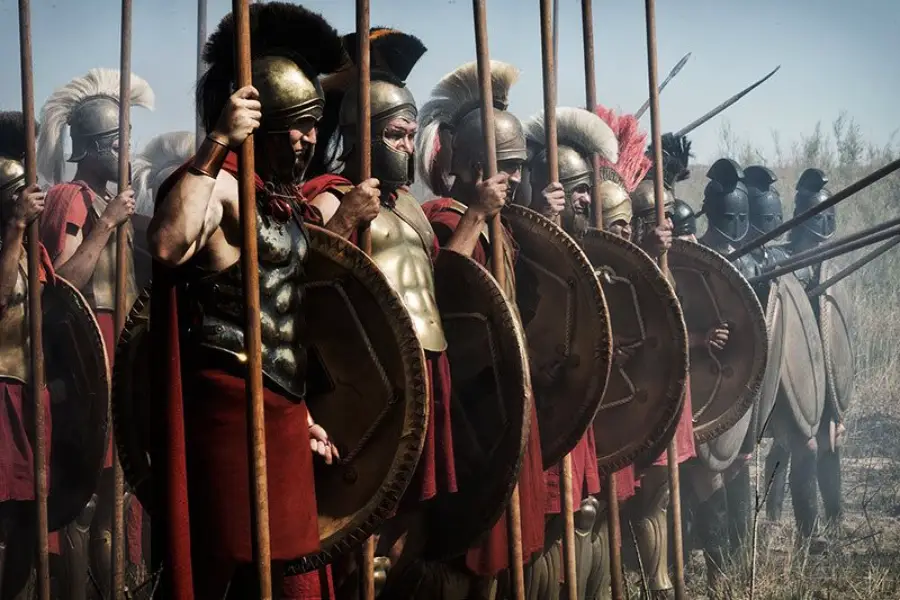 A group of ancient Greek soldiers in armor and helmets stand in formation, holding large round shields and long spears, ready for battle.