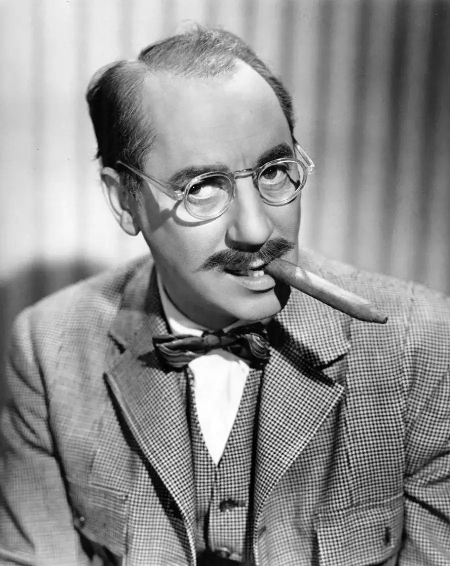 A man with glasses, a thick mustache, and bushy eyebrows wears a suit and bow tie while holding a cigar in his mouth, looking playfully at the camera.