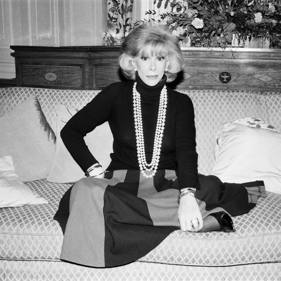 A woman with a voluminous hairstyle sits on a patterned sofa, wearing a long necklace, black turtleneck, and a long skirt. She faces the camera, with flowers and cushions visible in the background.