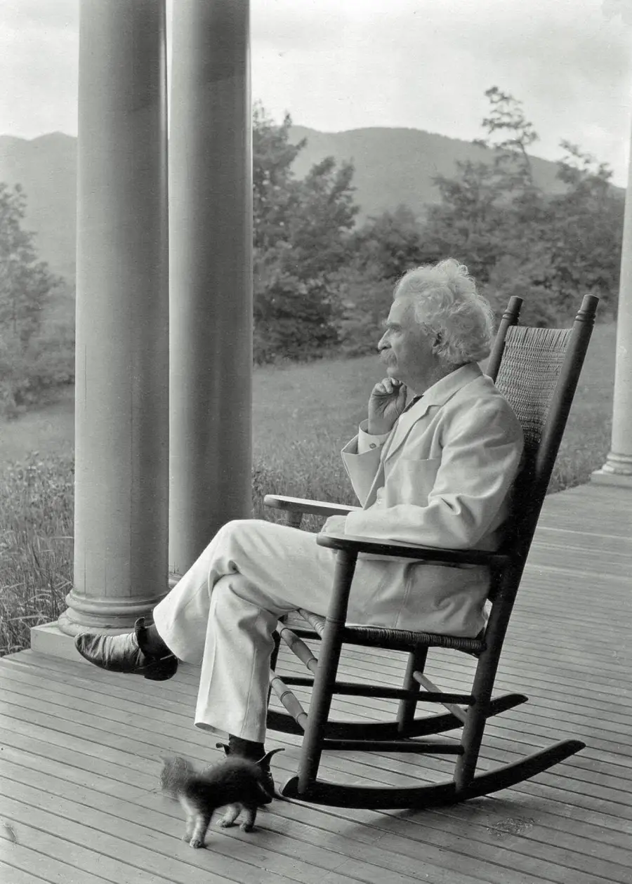 An elderly man in a white suit sits in a rocking chair on a porch, gazing thoughtfully into the distance, with trees and hills in the background and a small dog near his feet.