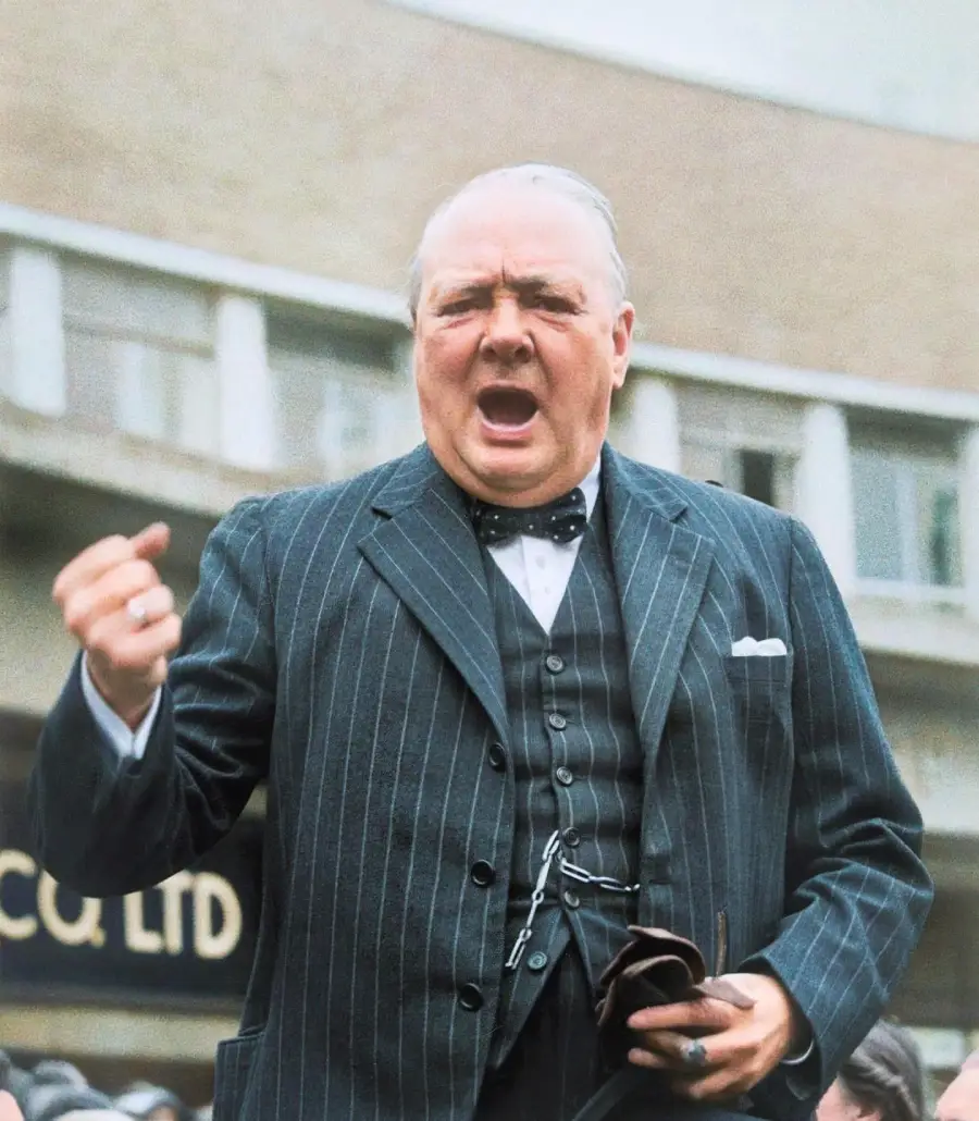 An older man in a striped suit and bow tie passionately speaks outdoors, raising his fist. The background shows a large building with windows and some signage.