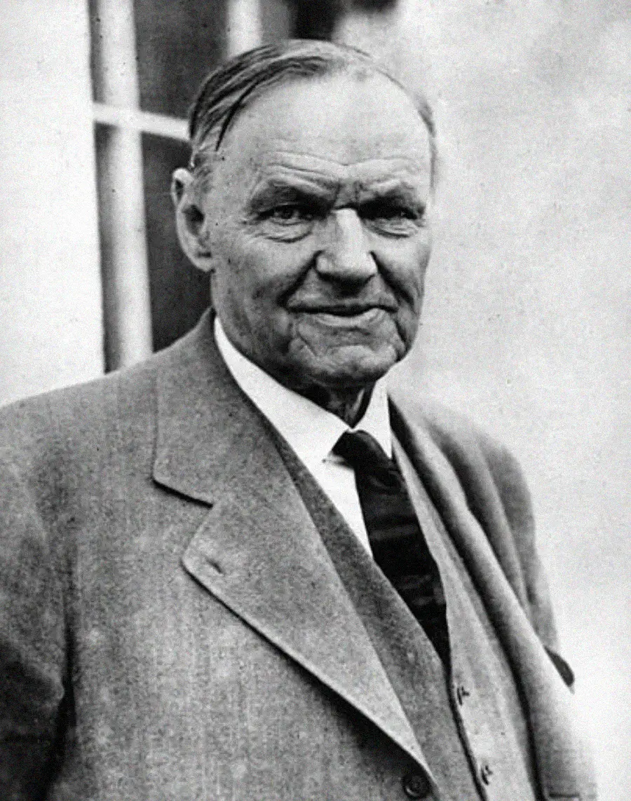 An older man in a suit and tie stands outside, looking at the camera with a slight smile. He has combed-back hair and a weathered face. The image is black and white.