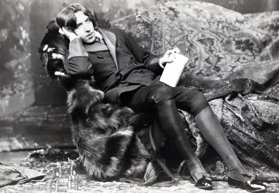 A person in vintage clothing reclines on a fur-covered chair, holding a piece of paper, with one hand resting against their head. Ornate rugs and patterned fabrics surround them. The photo is in black and white.