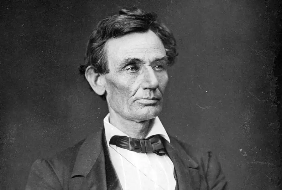 A black-and-white portrait of a man with dark hair wearing a suit, white shirt, and bow tie, looking slightly to the side with a serious expression against a plain background.