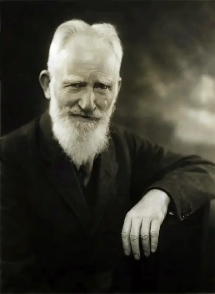 An older man with a white beard and mustache, wearing a dark suit, sits with one arm resting on a surface, looking directly at the camera and smiling slightly. The background is dark and blurred.