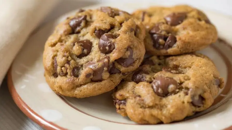Three chocolate chip cookies on a beige plate with a brown rim, sitting on a light-colored surface. The cookies appear soft and are generously studded with chocolate chips.