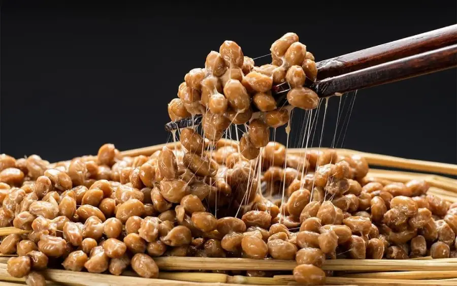 Chopsticks lifting sticky, stringy fermented soybeans (natto) from a woven basket against a black background.
