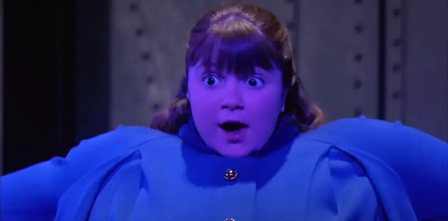 A girl with brown hair and bangs, wearing a blue outfit, looks surprised as her cheeks are puffed and her body appears swollen. The lighting gives her skin a purple-blue tint.