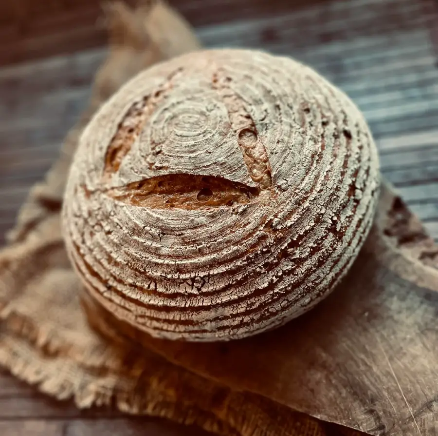 A round loaf of rustic bread with a spiral pattern and a single slit on top, resting on a piece of burlap atop a wooden surface.
