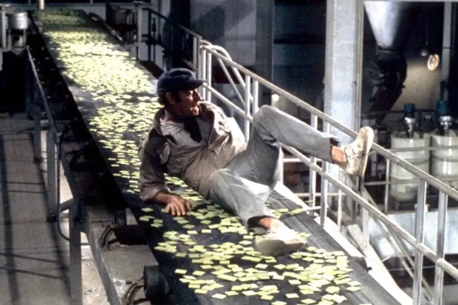 A man in casual work clothes and a cap sits on a conveyor belt covered with green rectangular objects inside an industrial factory setting.