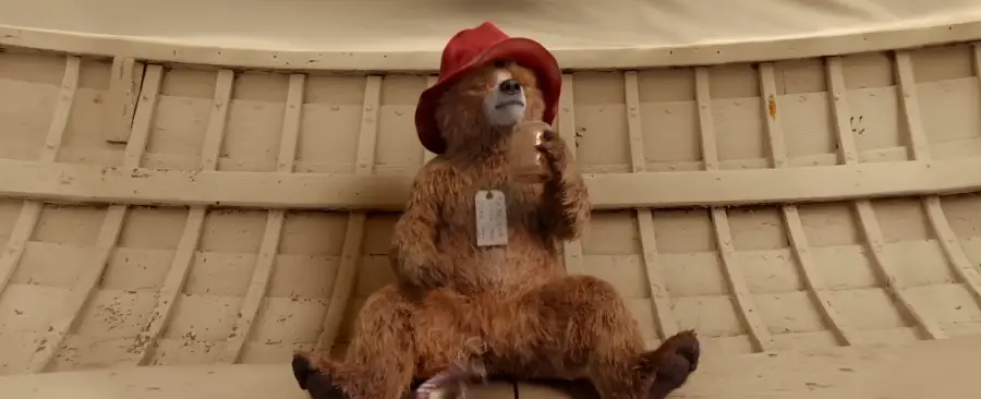 A stuffed bear with brown fur, wearing a red hat and holding a sandwich, sits against the curved wooden wall of a small boat. A tag hangs from its neck.