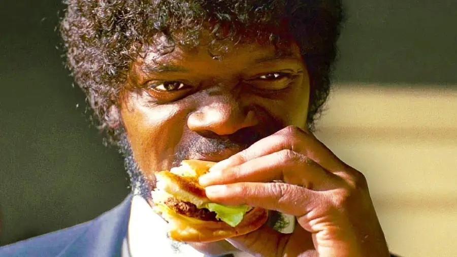 A man with curly hair and a beard, dressed in a suit, is staring intently while eating a hamburger, holding it close to his mouth.