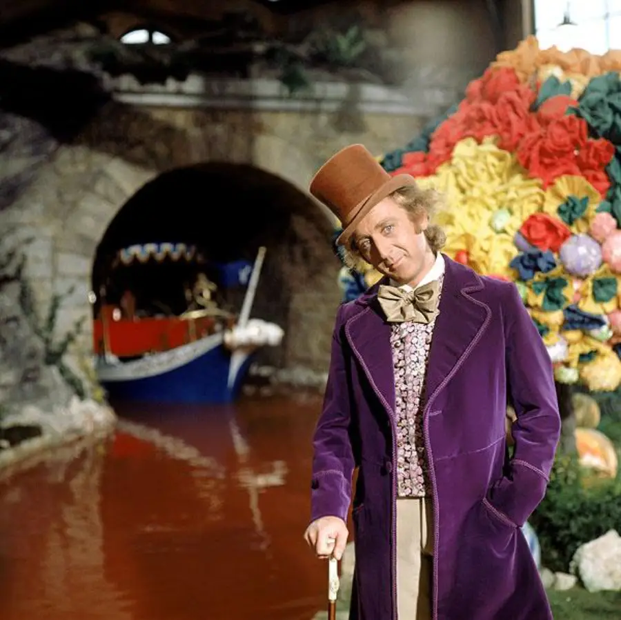 A man in a purple velvet coat and brown top hat stands by a candy-filled riverbank, holding a cane. In the background, a boat floats on a chocolate-colored river under a stone bridge. Large colorful candies decorate the scene.