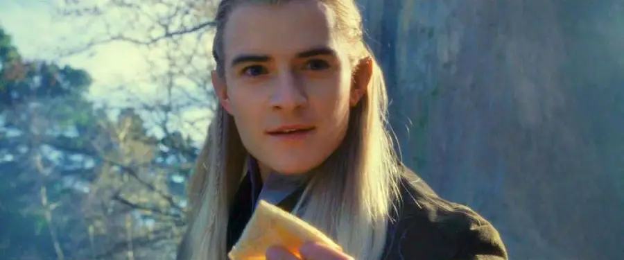 A person with long, straight, blonde hair and pointed ears stands outdoors, holding a piece of food and looking forward with a slight smile. Trees and a stone wall are visible in the background.
