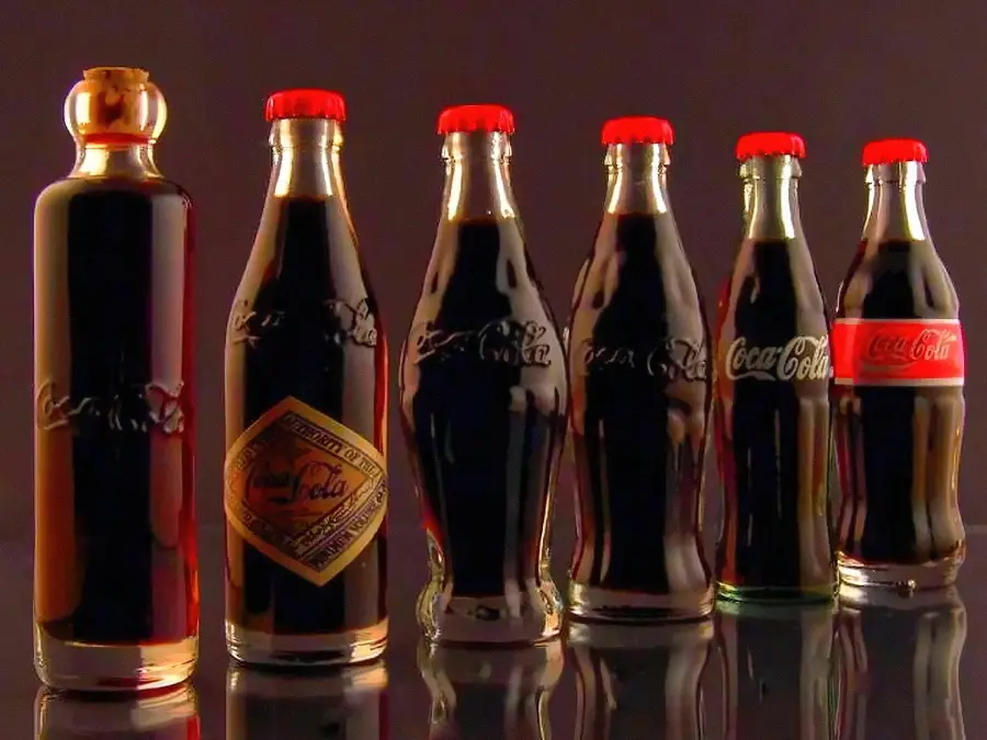 Six Coca-Cola bottles in a row, each with a different shape and label design, showing the evolution of the bottle from left to right, all filled with dark soda and topped with red caps against a dark background.