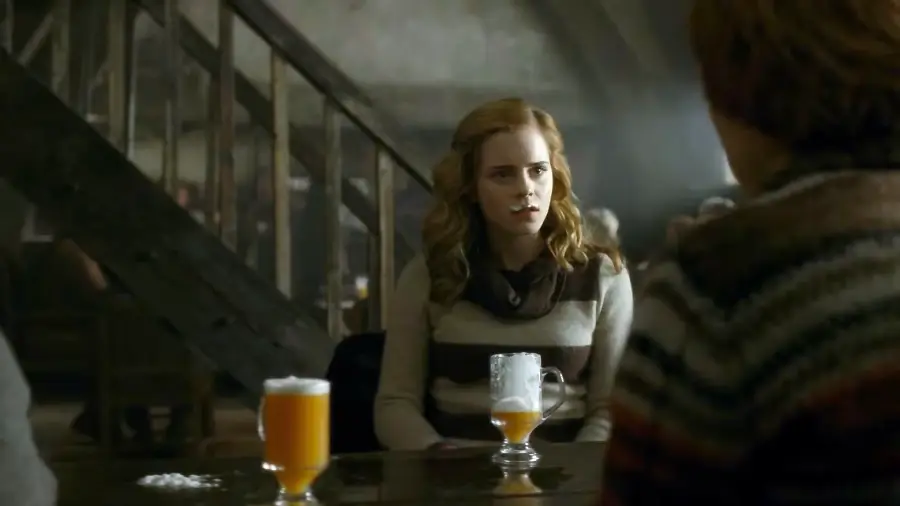 A young woman with wavy blonde hair sits at a wooden table in a dimly lit room, facing another person. Two frothy drinks are on the table. A staircase and blurred figures are visible in the background.