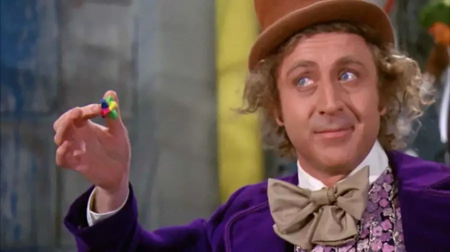 A man in a purple velvet coat and brown top hat holds up a colorful candy while smiling. He has curly blonde hair and wears a large tan bow tie over a patterned vest. The background is slightly blurred.