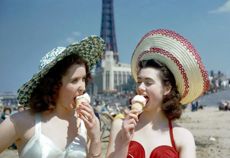 Two women in sun hats and swimsuits eat ice cream cones on a sunny beach, with a tall tower structure and crowds visible in the background.